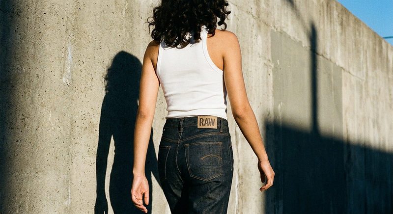 Back view of a person in a white tank top and dark denim against a sunlit concrete wall