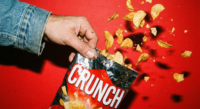 Hand grabbing a chip from a red snack bag with chips scattered on a red backdrop